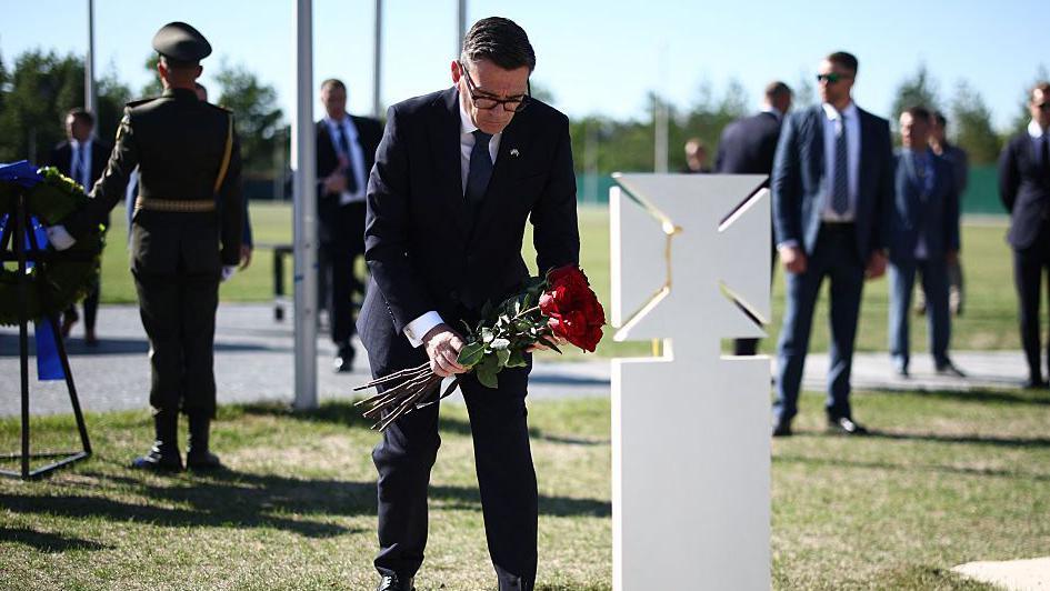 Burnham bends down holding a bunch of red roses in front of a white gravestone as people stand behind him
