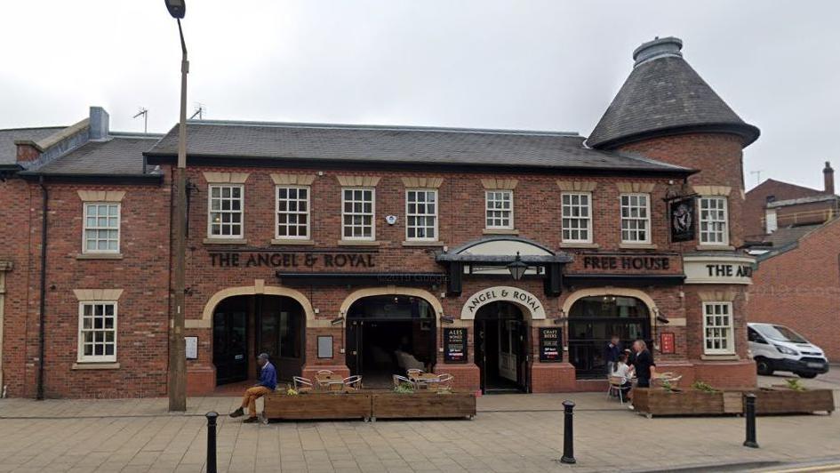 A red brick pub with the lettering "The Angel & Royal Free House" above the four entrance arches. Several customers can be seen sitting in chairs on the right.  A man sits on a planter on the left with his legs crossed.