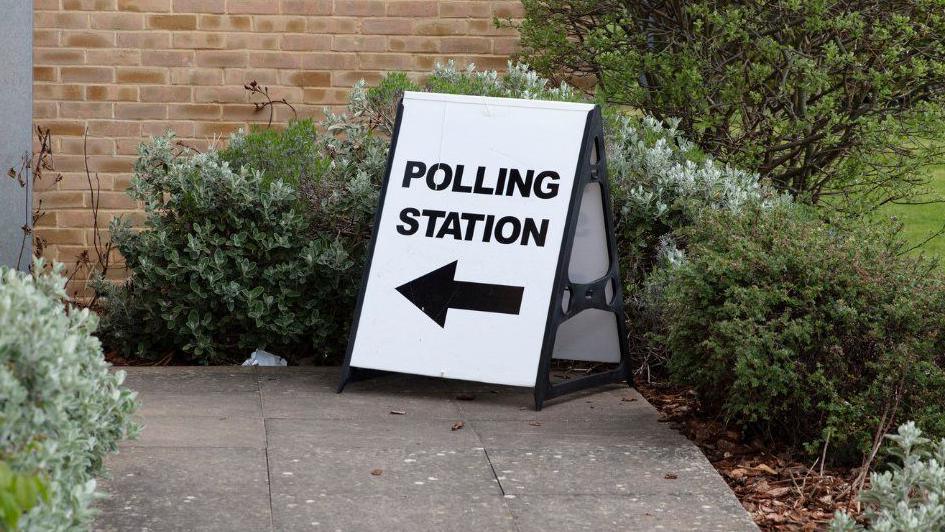 A polling station sign, with POLLING STATION, written on it in capital black letters, with an arrow to the left, outside a building with folliage