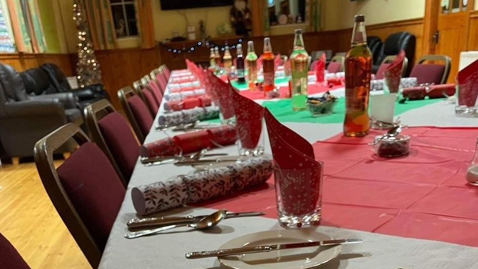 A table set for a community Christmas Day dinner in Ardess Parish Church hall in Kesh in December 2023.  The long table is laid with white, green and red table cloths, plates, glasses, cutlery, crackers and bottles of grape juice.