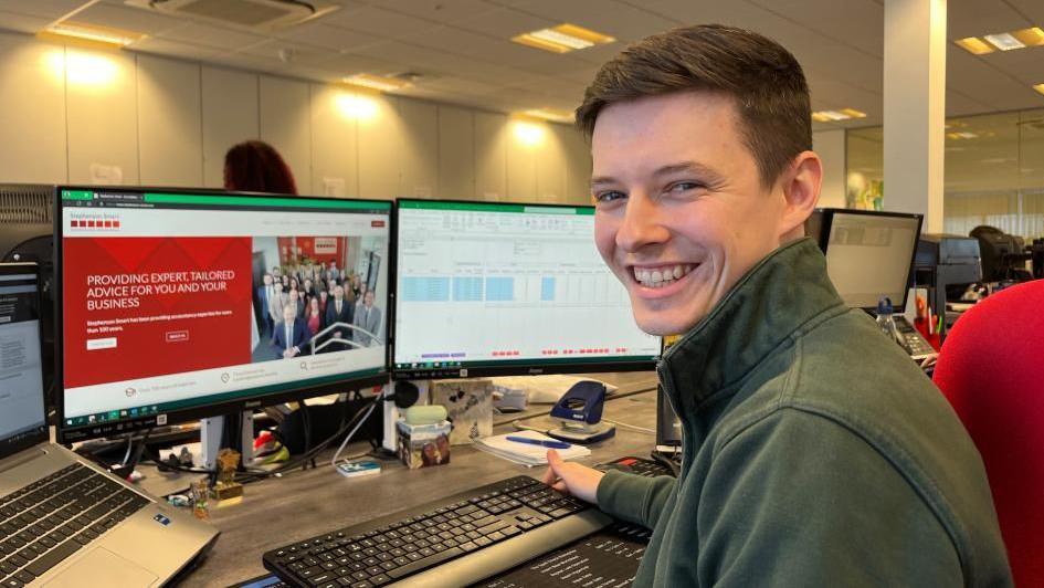 Connor Simpson is wearing a green jacket and is sitting on a red office chair. He has short brown hair, and is smiling, looking at the camera. In front of him is a three-screen computer displaying work-based websites and forms.