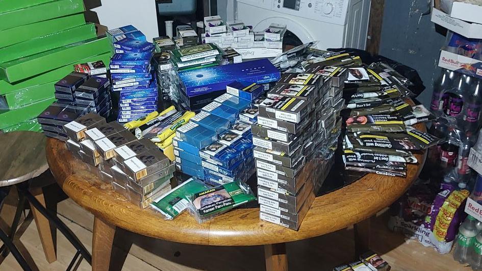 A large pile of illegal cigarettes and tobacco stacked on a round table in a dimly lit room.