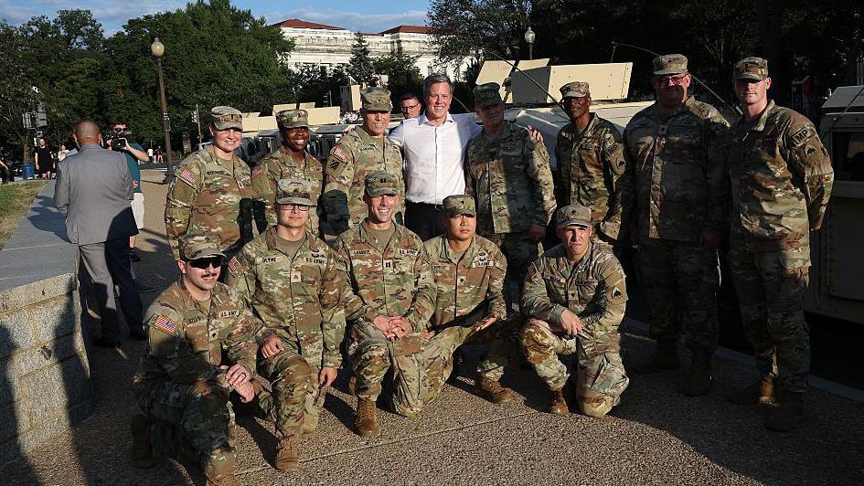 A row of National Guardsmen in camouflage uniform kneel in front of a row of standing guards, with Dan Driscoll in white button-down shirt in the middle with his arms around two of them