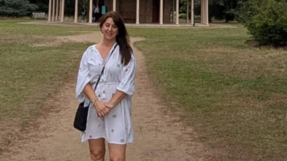 A picture of a woman in a park. She is wearing a white summer dress and has a black clutch bag on her shoulder. She has long dark brown hair which she's let down on one side, but is behind the ear on the other side.