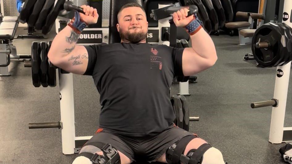A man with dark hair and a beard in a black t-shirt and shorts is in a gym, lifting very large weights. There are large straps and supports around his legs