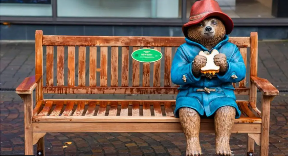 A statue of a Paddington Bear in a red hat and a blue coat holding a sandwich and sitting on a bench