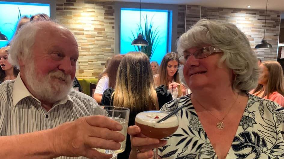 A man and a woman cheers their glasses, and look into each others eyes. The man, Ian, has grey hair, a grey beard and is wearing a stripy shirt. Vera has grey hair, glasses and is wearing a black and white top. They are smiling at each other.