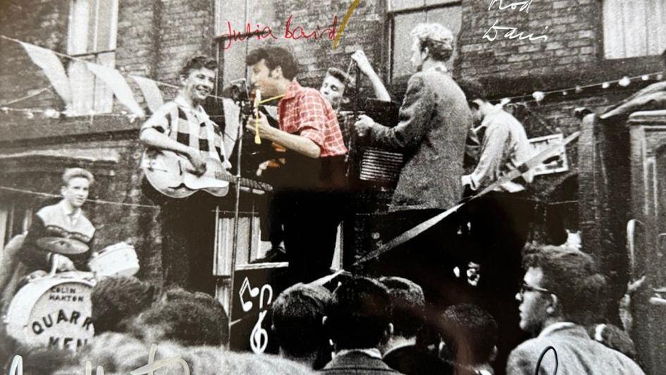 A black and white photo of The Quarrymen playing in front of a house. There is a drummer and multiple guitar players in the six-strong group. A young John lennon sings into a microphone. The image has been changed to colourise him. He has short brown hair and wears a red check shirt.