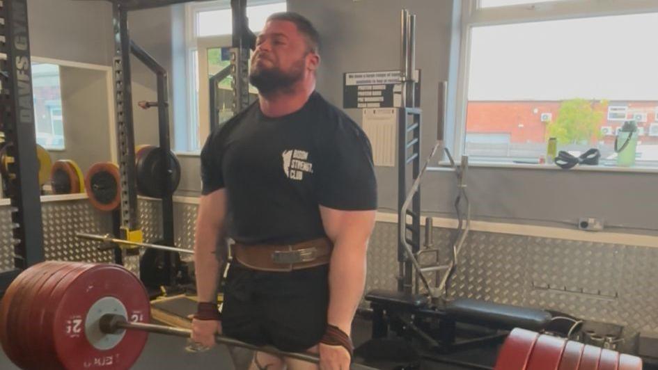A man with dark hair and a short beard is straining as he lifts very large weights suspended either side of a metal bar