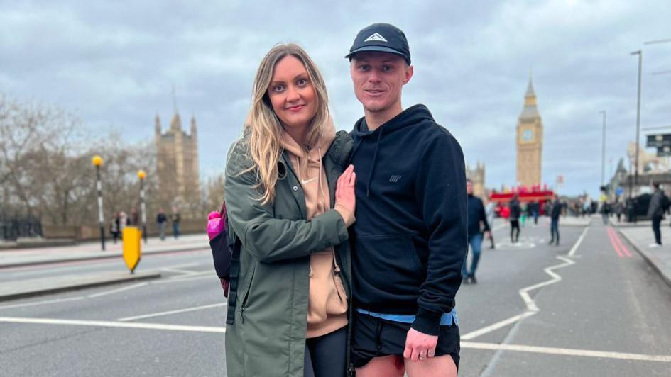 Jack Taylor and his wife, Ellen, after the half marathon last year. They are standing on Westminster bridge with Big Ben in the background. Ellen has blonde hair past her shoulders and wears a dark green, long coat over a beige jumper and dark blue pants. Jack has a dark blue baseball cap on,a dark blue top and dark shorts.