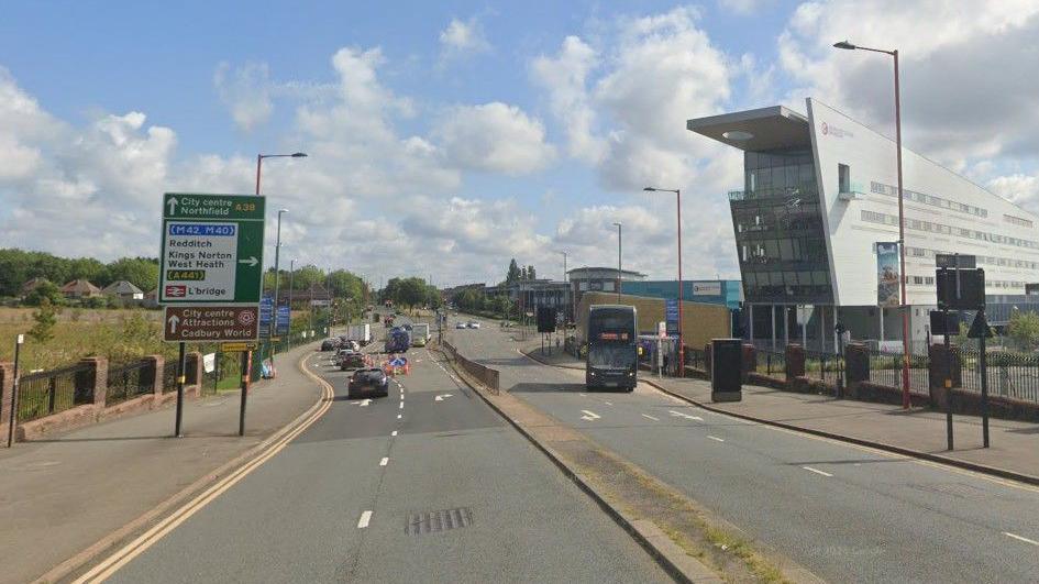 A dual carriageway with cars on one sign, a green and white sign for directions and a bus on the other side. A metal railing goes down the central reservation.