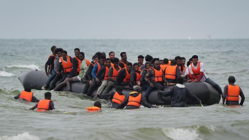 A large group sitting on an inflatable small boat, many sitting astride the outer edges. Six people are seen wading in deep sea water towards the boat. Most, but not all, are wearing orange life-jackets.