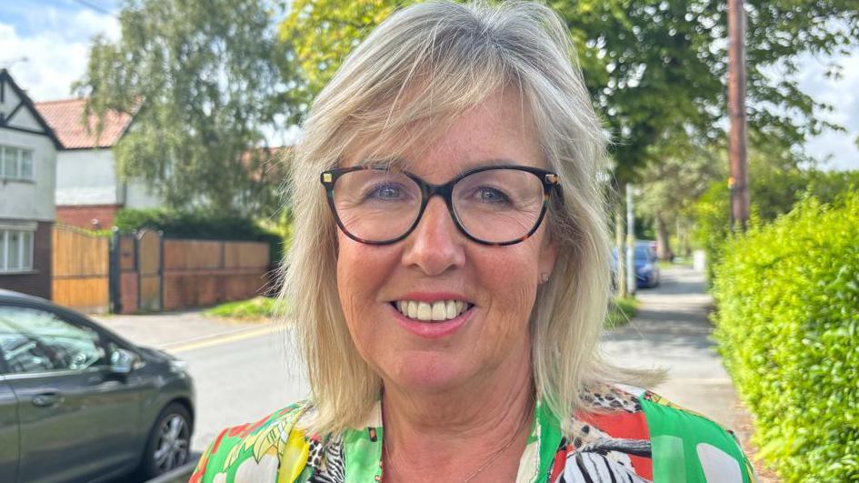 A lady with short blonde hair and glasses is pictured on a street.
Behind her left shoulder is a green hedge and behind her right shoulder is a road with houses in the background. 