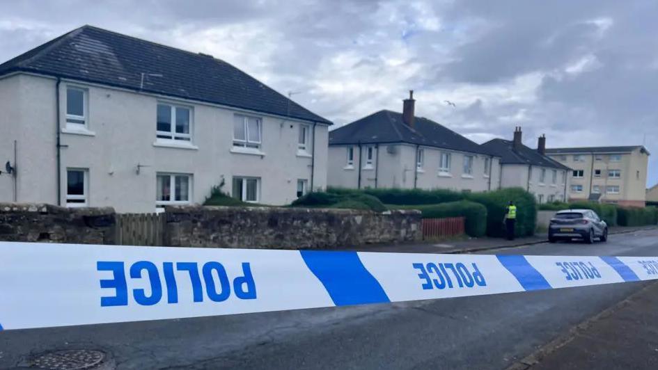 Blue and white police tape which is upside down in front of a row of cream two-storey buildings. A police officer is visible in a distance.