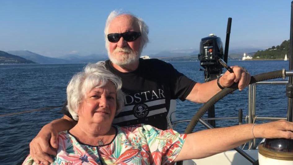 Two people, Ian and Vera, sit on a boat looking at the camera. Ian has white hair and a white beard and is wearing sunglasses. Vera wears a colourful patterned top. The sea is navy and the sky a cloudless blue. 