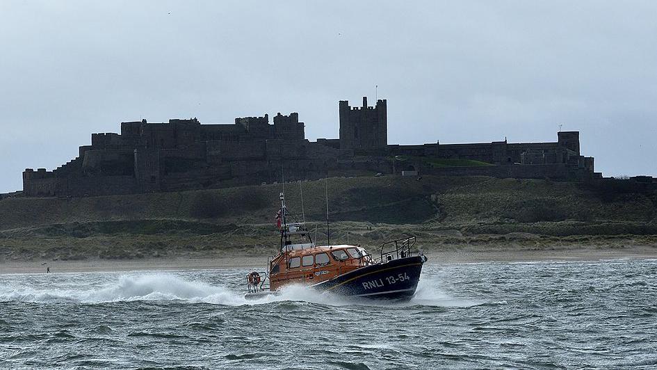 The blue and orange RNLI lifeboat in the water in front of Bamburgh Castle.