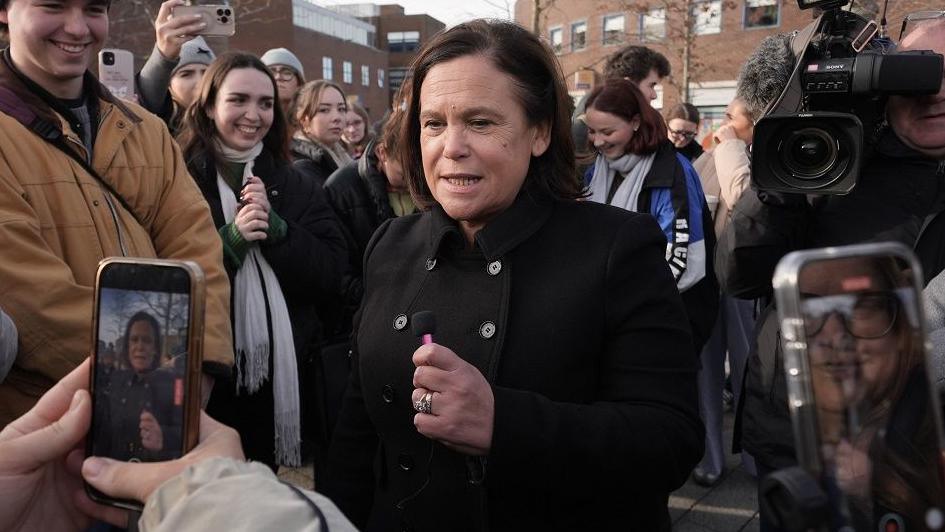 Mary Lou McDonald wearing a black button-up coat at an election event in Dublin.  Two people are taking pictures of her with their phones while a cameraman is behind her. 