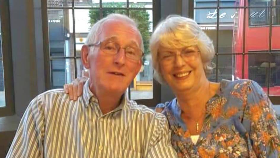 A selfie style shot of John Barrie, a man in his 70s with white hair. His wife Jeanette is next to him wearing black glasses and smiling at the camera.
