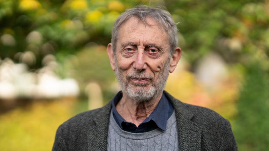 Michael Rosen in a grey jumper and grey coat front of a green background that has been blurred.