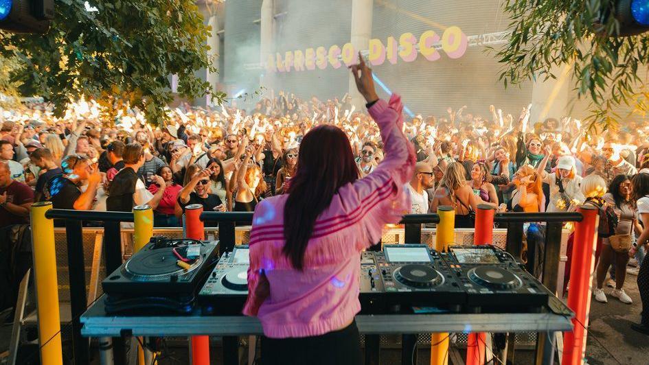 A DJ waves her arms in front of a large crowd dancing at an Alfresco Disco event in Bristol