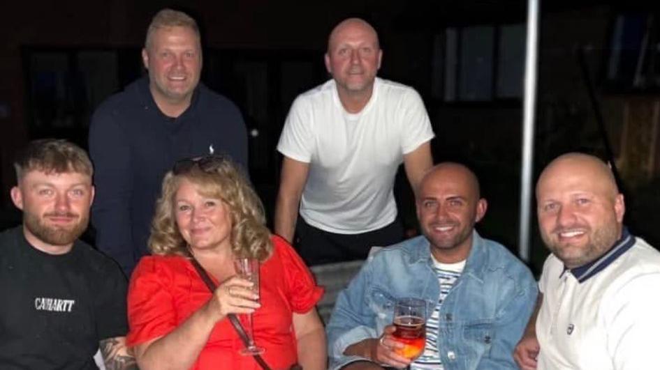 A group photo shows a woman in a red dress surrounded by five men. Three of the men are sitting on either side of her while two are standing beside her. They are all smiling.