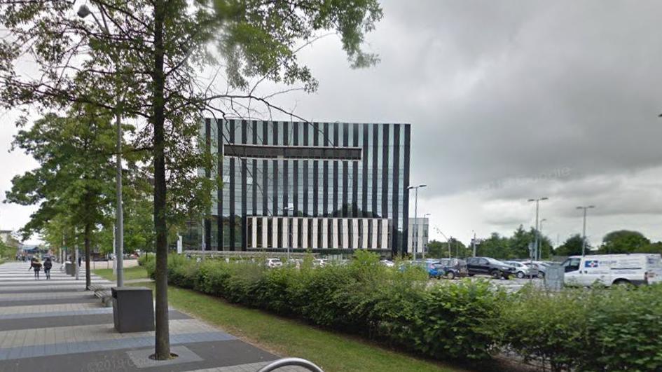 Large metal and glass civic building with a car park in the foreground and a tree-lined path to the right