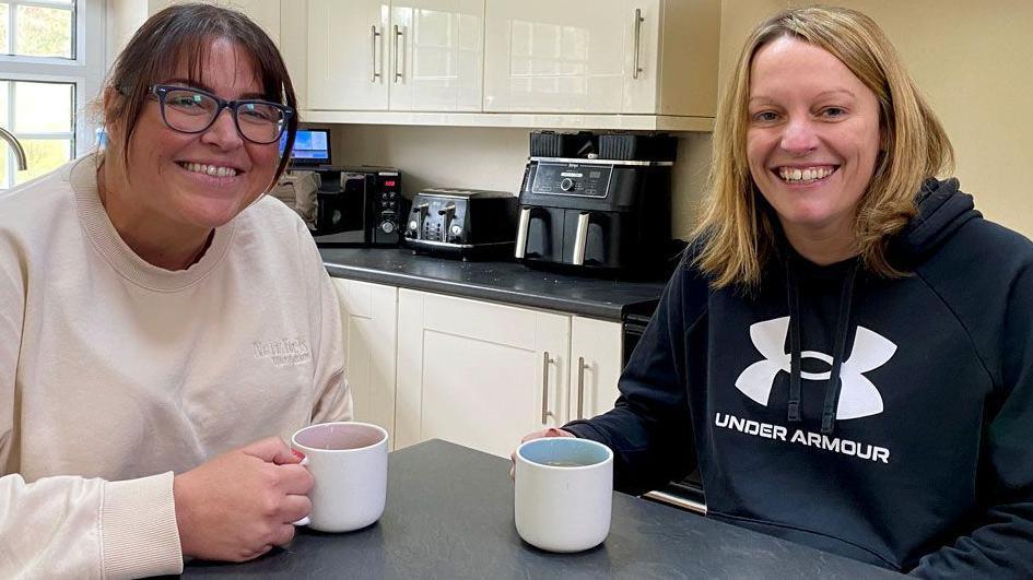 Jaycee Charlton and Katherine Singer who are both in their late 30s are sitting at a farmhouse kitchen table drinking cups of tea and smiling. Katherine has blonde shoulder-length hair and a blue Under Armour top. Jaycee, who has glasses and dark hair, is wearing a white top