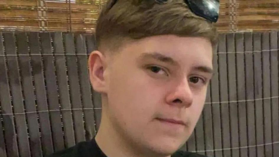 A teenage boy with dark hair and sunglasses on his head. He is starting into the camera. A fence is behind him.