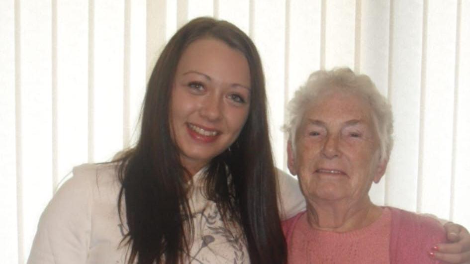 Rachael is stood with arm around her gran Lilian. Rachael has long brown hair, she is wearing a white hoodie with a motif on the front. Lilian is wearing a pink knitted jumper, she has short grey hair. 