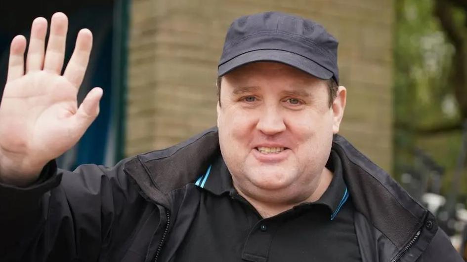 Peter Kay wears a dark cap and black t-shirt and coat and waves at the camera.