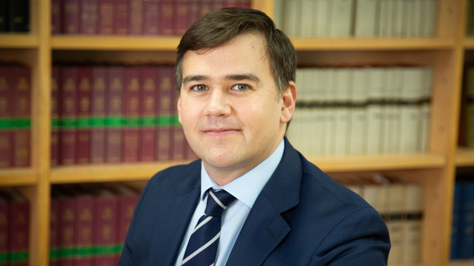 A middle aged man looks towards the camera with a neutral face. He has dark hair and is wearing a light blue shirt with a navy jacket and tie. He is in front of a bookshelf holding various leather bound books.