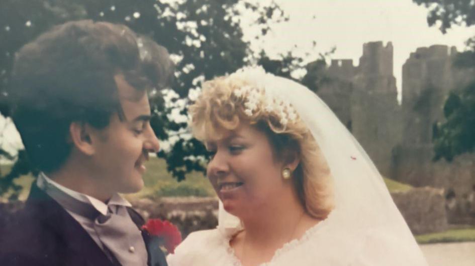 Trasi and Mark Coghlan smile at each other, Trasi is wearing a white veil and dress. She has long blond hair. Mark is wearing a black suit jacket with a grey vest and white collar. He has a moustache and brown short hair. Behind the couple a castle is visible between the trees.