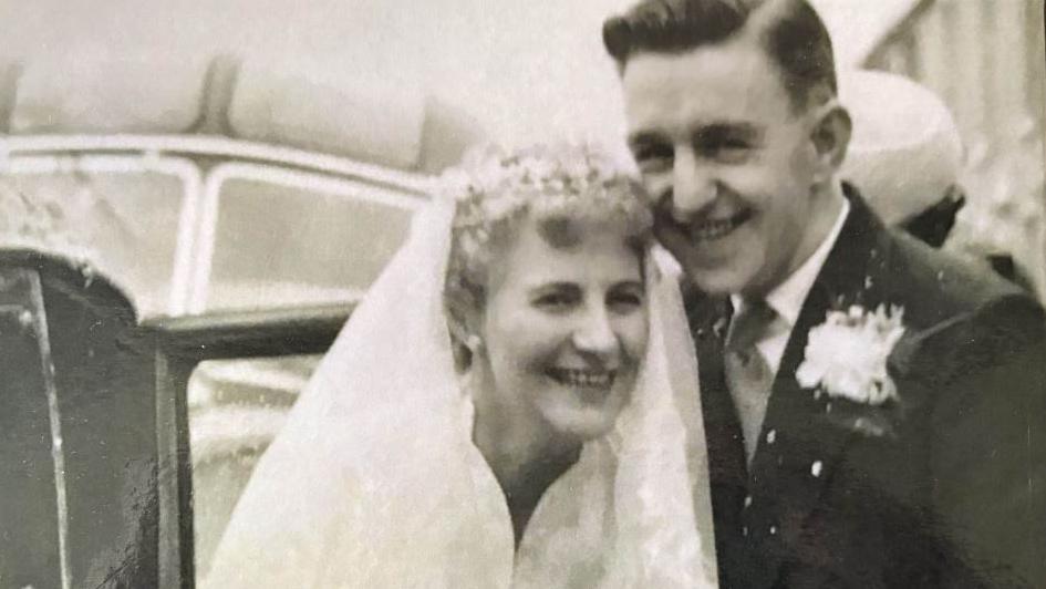 Gordon and Joan Cilgram on their wedding day in 1958 smiling standing in front of a wedding car. Joan with short blond hair wears a white veil and wedding dress and Gordon wears a black suit with a boutonnière 