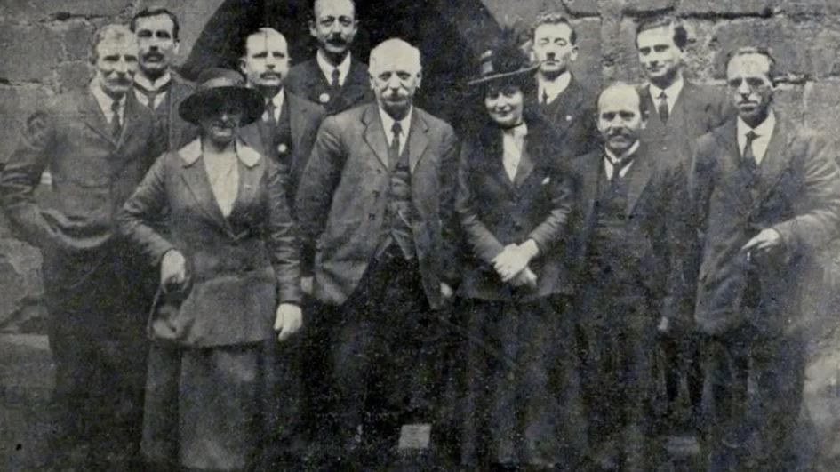 A black and white photograph and a group of men and women stood in front of  a building.