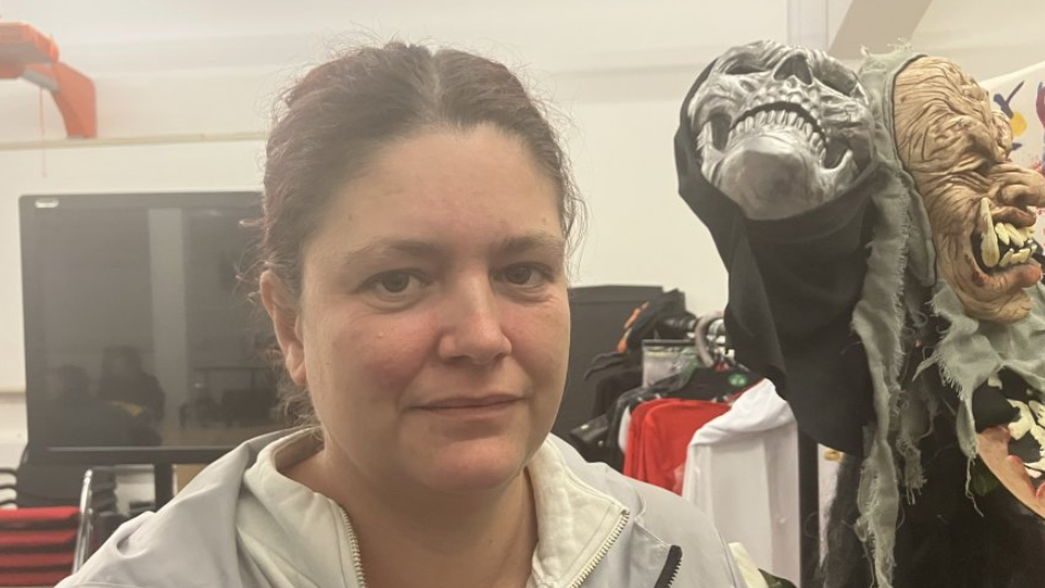 Woman with brown hair tied back wearing a white jacket. She is standing next to some Halloween masks. One is a grey skeleton in a black hood and the other is a monster with very big teeth