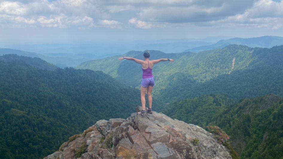 Francesca Murphy stands on top of a mountain looking at a beautiful view of green, lush mountains. Her back is to the camera and she is holding her arms out and looking at the view. 