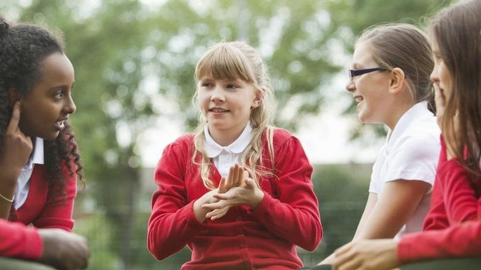 Four secondary school pupils talking to each other wearing uniforms of white T-shirts and red jumpers  with a blurred trees in the background