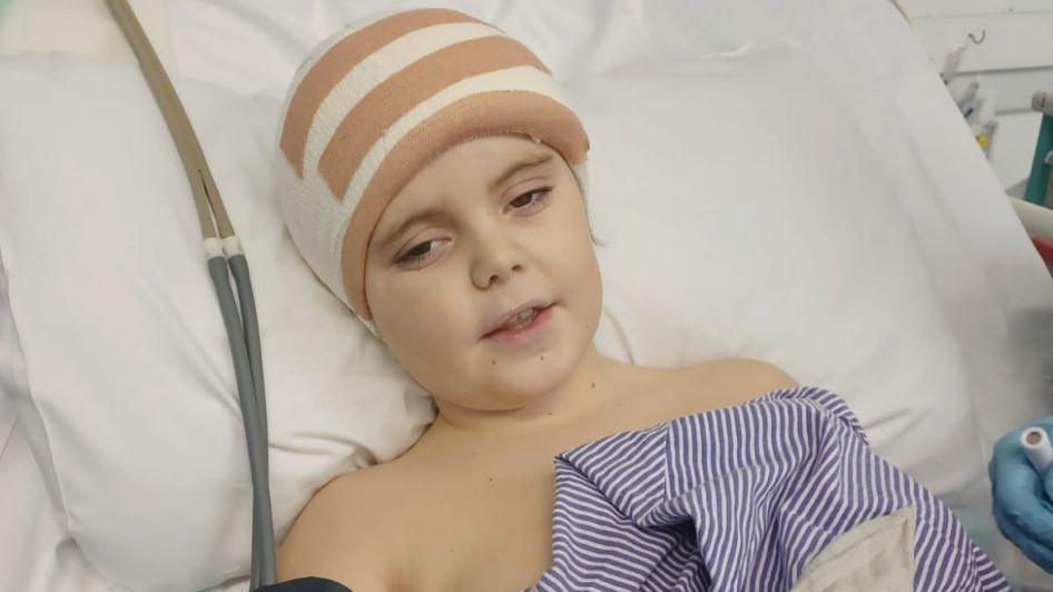 A young boy lying in a hospital bed with a large bandage on his head.