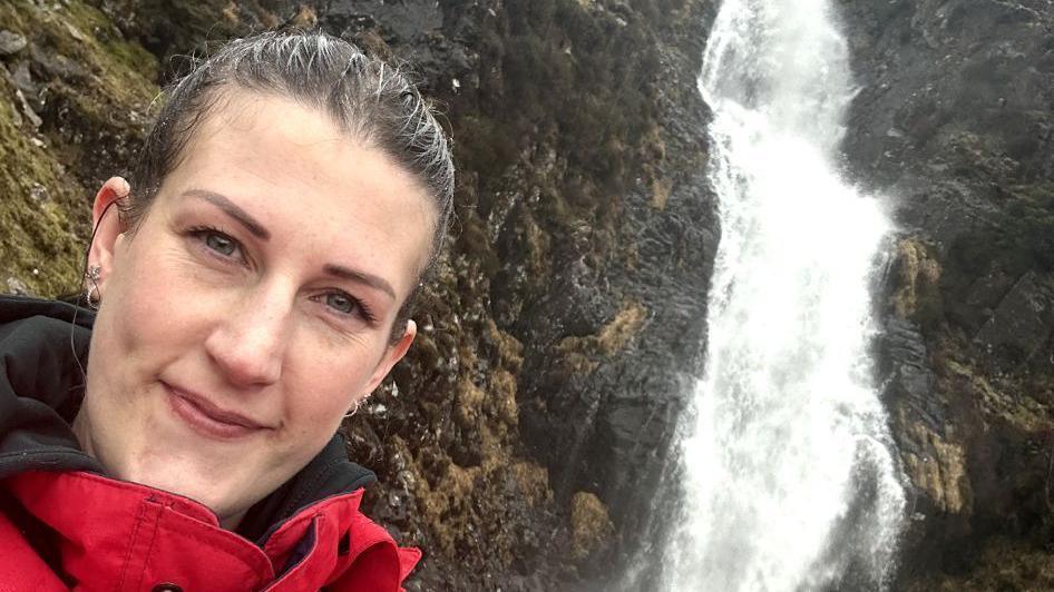 Ruth Trinder standing in front of a waterfall. Her dark hair is pulled back behind her head, she has small hoops in her ears and is giving a slight smile to the camera. She wears a red and black jacket