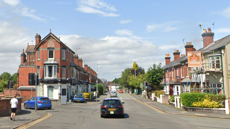 A crossroads with traffic lights on a sunny day. There are terraced brick houses on either side of the street running straight ahead. There is a person in a white t-shirt and black shorts walking along the street.