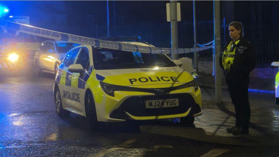 A female police officer stands next to a police car next to police tape connected between two lampposts, with other emergency vehicles seen in the background. 