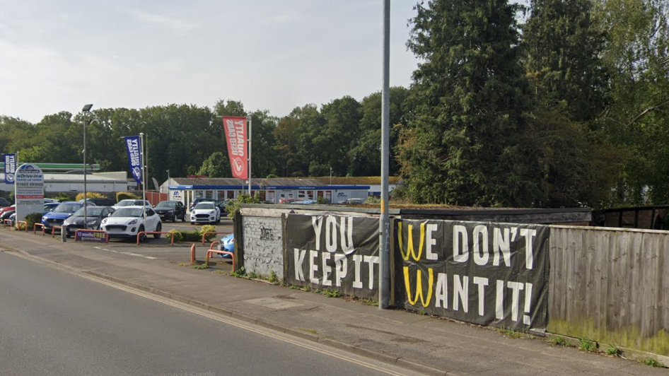 A sign hanging on a fence that reads "You Keep It. We Don't Want It!". Behind the sign is a car park.  