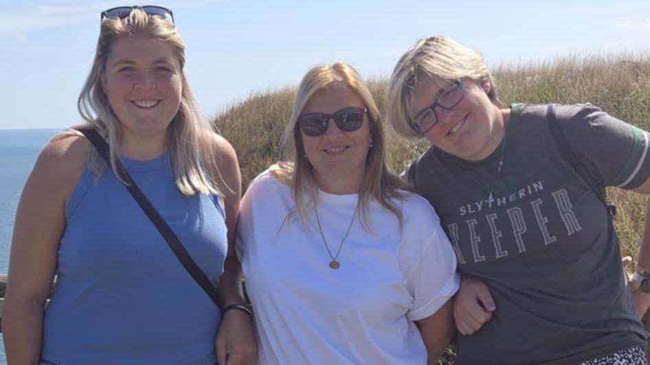 The photograph shows three blonde women at the seaside. The woman on the left wears a blue top and has a bag slung around her chest. A pair of sunglasses sit on top of her head, keeping her hair out of her face. The woman next to her wears a white t-shirt, a gold necklace and sunglasses while the woman on the very right is wearing a grey t-shirt that reads "Slytherin Keeper". She is wearing glasses.
