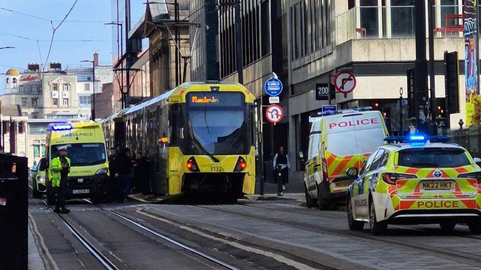 A stationary yellow and black Metrolink tram, flanked by an ambulance and police vehicles, at the scene of the collision.