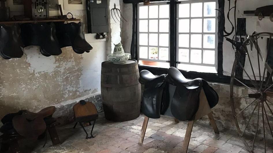 A bare room with brick and plater walls, old saddles are placed on a wrack and there is a large bicycle wheel on show