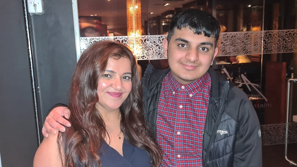 Pooja and her son Ronan are stood together outside a restaurant. Ronan has a black jacket on and checked shirt and has his arm around his mother who has a navy sleeveless top on.