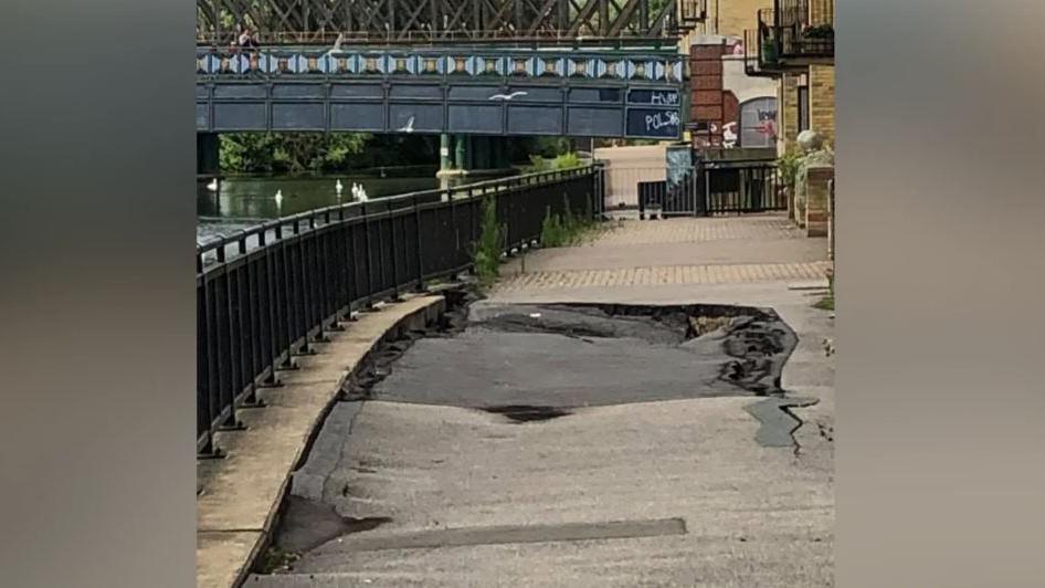 A massive sinkhole in the footpath along the River Nene with a bridge in the background.