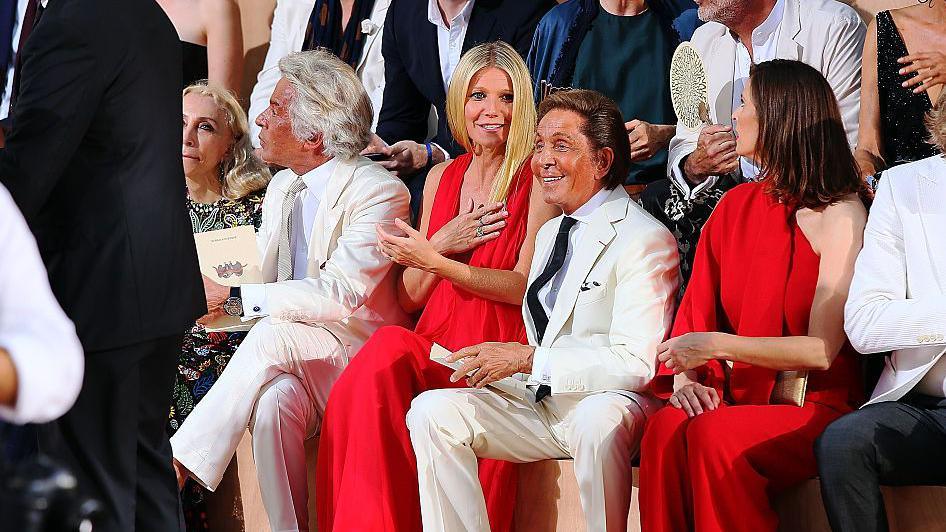 Giancarlo Giammetti, Gwyneth Paltrow dhe Valentino Garavani marrin pjesë në sfilatën e modës Valentino Mirabilia Romae në Piazza Mignanelli më 9 korrik 2015 në Romë, Itali. Giancarlo dhe Valentino janë ulur në të dyja anët e Paltrow, të dy të veshur me kostume të bardha. Paltrow, në mes, është duke buzëqeshur, duke mbajtur njërën dorë në gjoks, me Valentino-n e ulur pranë saj në rreshtin e parë të sfilatës duke buzëqeshur.