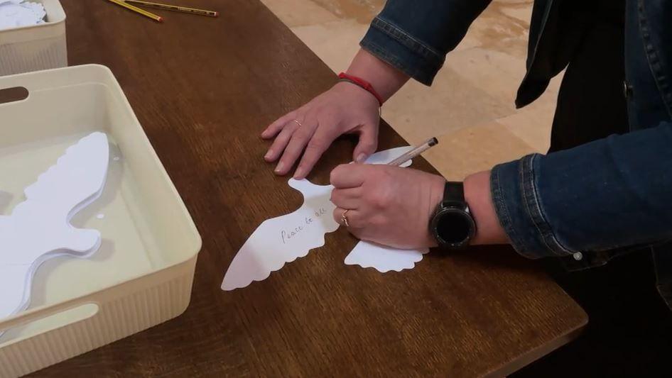 A white paper cut-out of a dove with someone writing "peace to all" on it with a box next to it containing more dove cutouts.