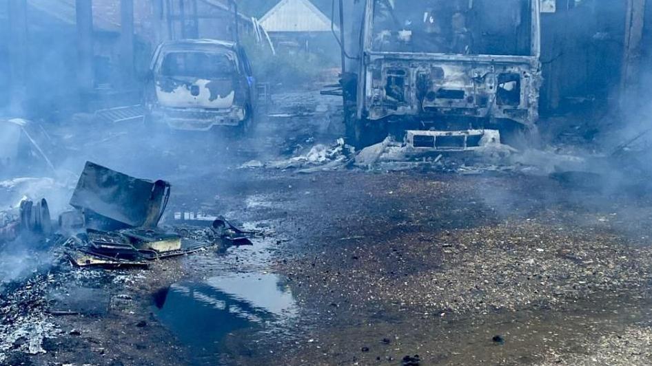 Two burnt out vehicles surrounded by smoke and water puddles. On the right is a lorry facing on, and on the right is the rear of a car. There is other blackened debris on the ground around them. They are between two burnt out barn structures.